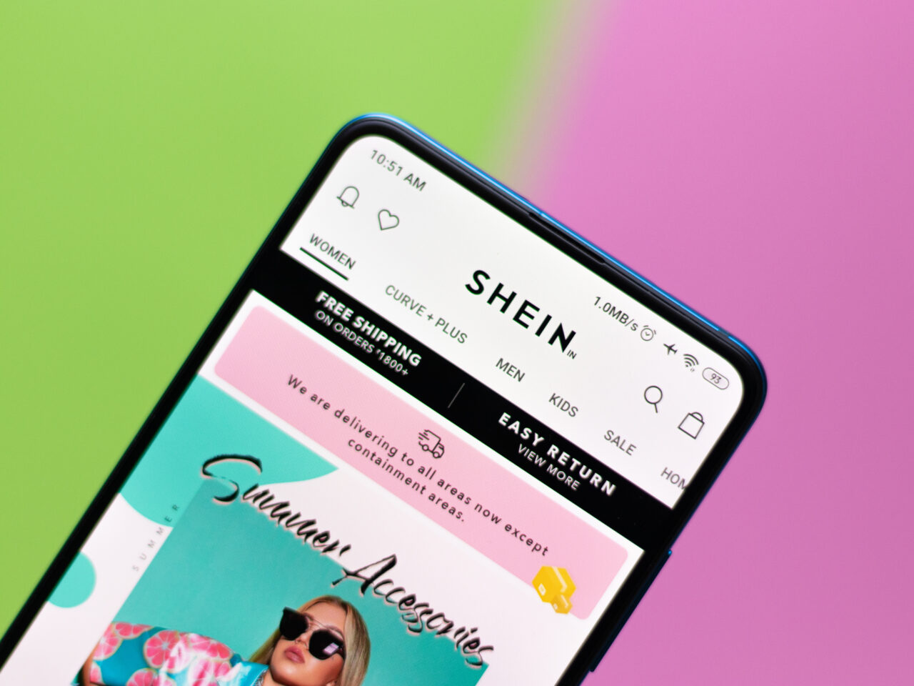 Shein opinie. Smartfon z otwartą aplikacją sklepu SHEIN na ekranie, wyświetlającym reklamę akcesoriów letnich z ikoną kobiety w okularach przeciwsłonecznych na kolorowym tle zielono-różowym.