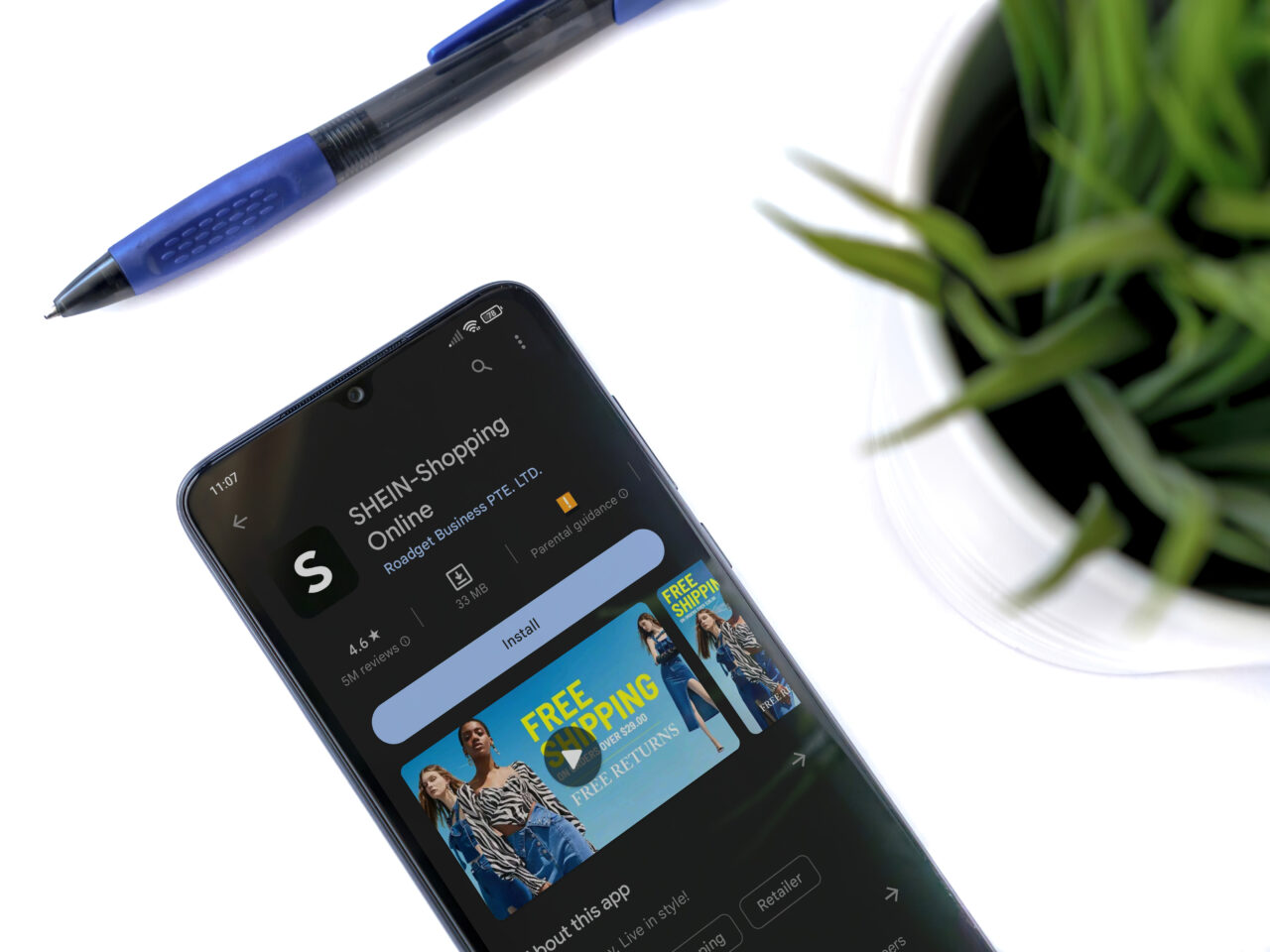 Smartfon wyświetlający stronę sklepu internetowego SHEIN na białym tle z niebieskim długopisem i doniczką z zielonymi roślinami.