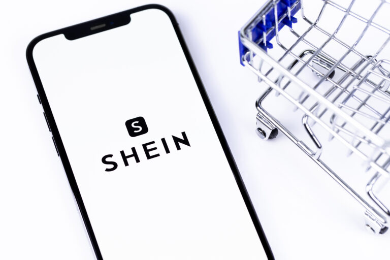 Smartfon z otwartą aplikacją Shein obok połowy metalowego koszyka na zakupy na białym tle.