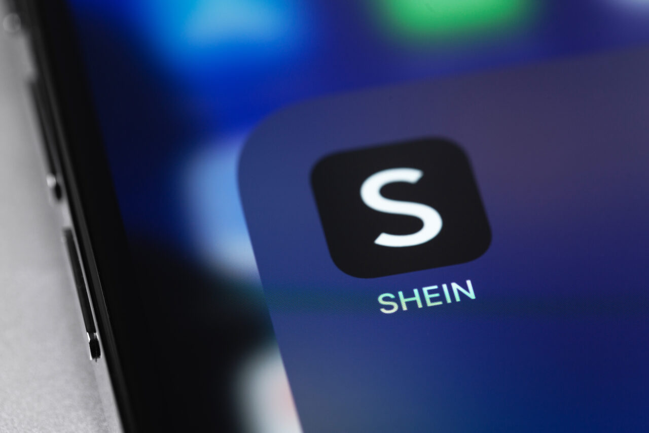 Ikona aplikacji SHEIN na ekranie smartfona z rozmytym tłem w odcieniach niebieskiego.