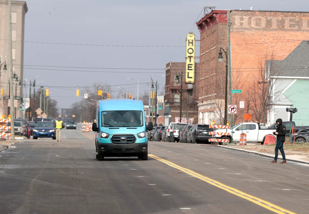 Niebieski samochód dostawczy jedzie ulicą miejską, w tle stary budynek z neonem HOTEL oraz osoba z kamerą filmująca pojazd. Droga zawiera znaki drogowe i barierki ostrzegawcze wskazujące na prace drogowe.
