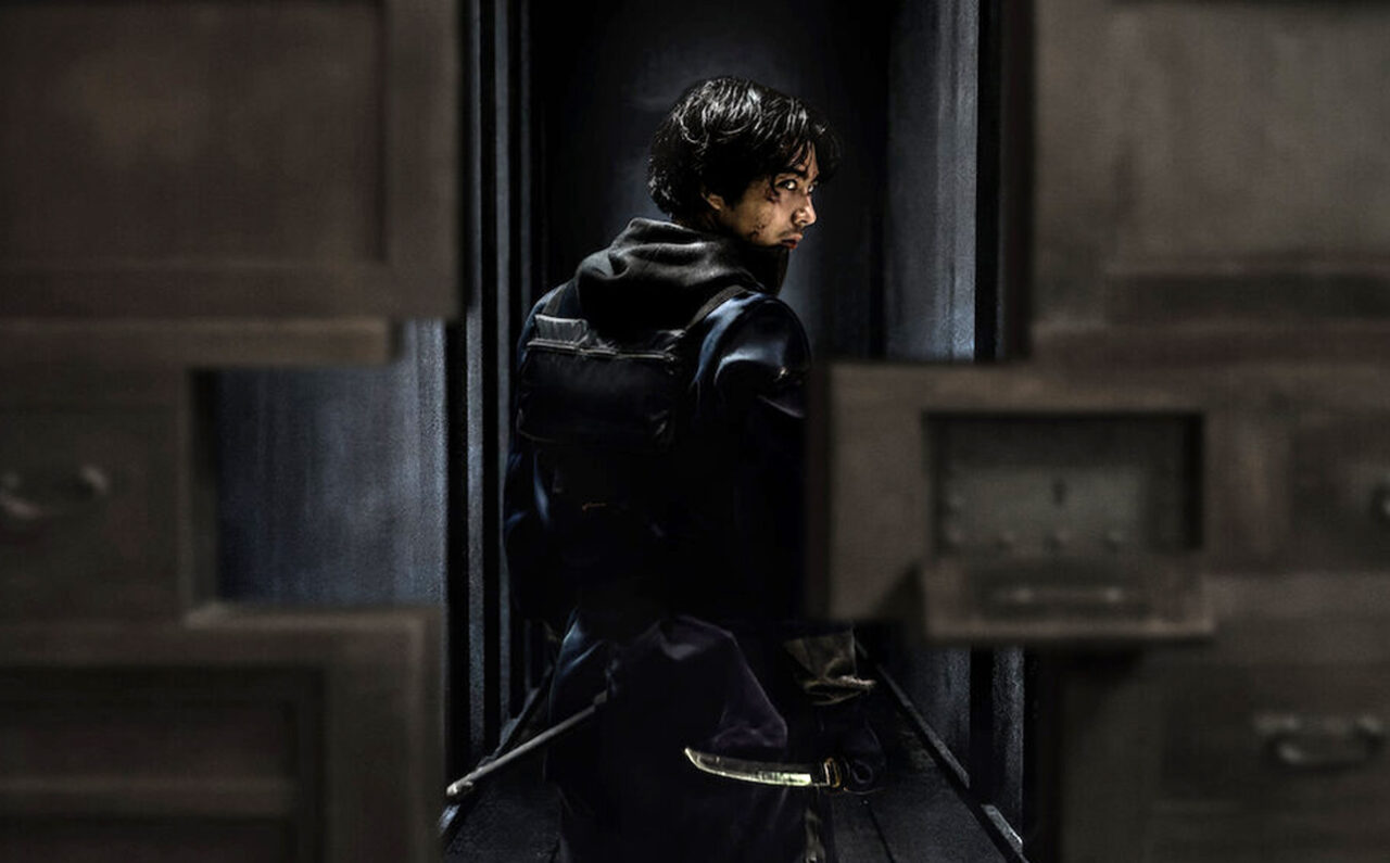Mężczyzna w czarnym stroju i z mieczem, spoglądający przez ramię, stojący w ciemnym przejściu między otwartymi drewnianymi drzwiami.