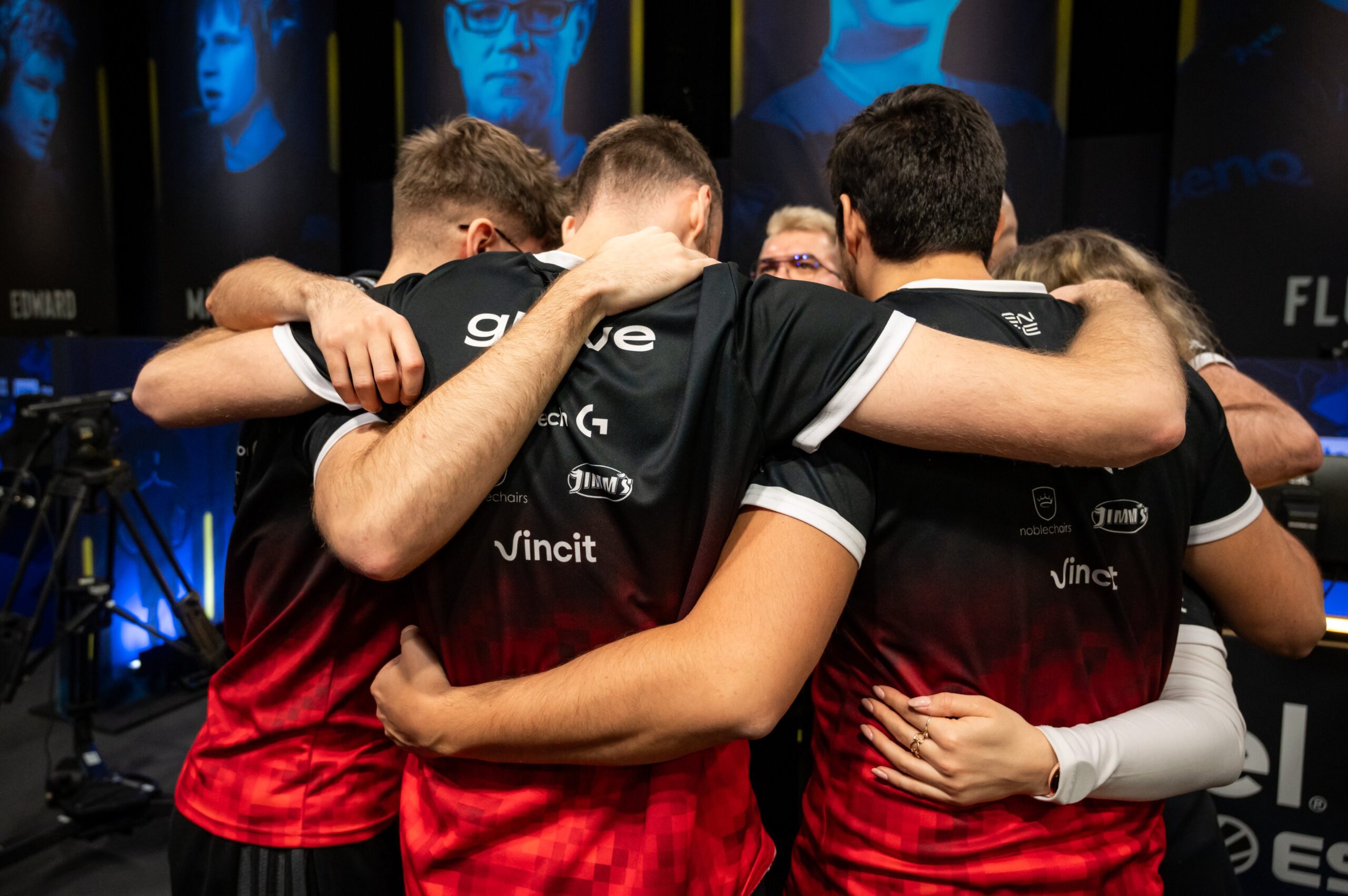 Grupa graczy e-sportowych na IEM Katowice 2024 w czarno-czerwonych koszulkach przytulająca się i tworząca krąg, ze skupionymi wyrazami twarzy, na tle rozmytych scen z ich wizerunkami.