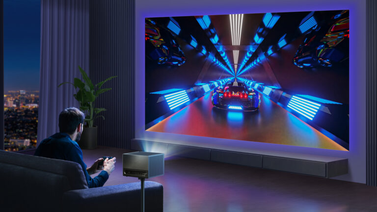 Mężczyzna siedzi na kanapie w pokoju z widokiem na miejskie nocne światła i gra w wyścigową grę wideo na dużym ekranie telewizora.
