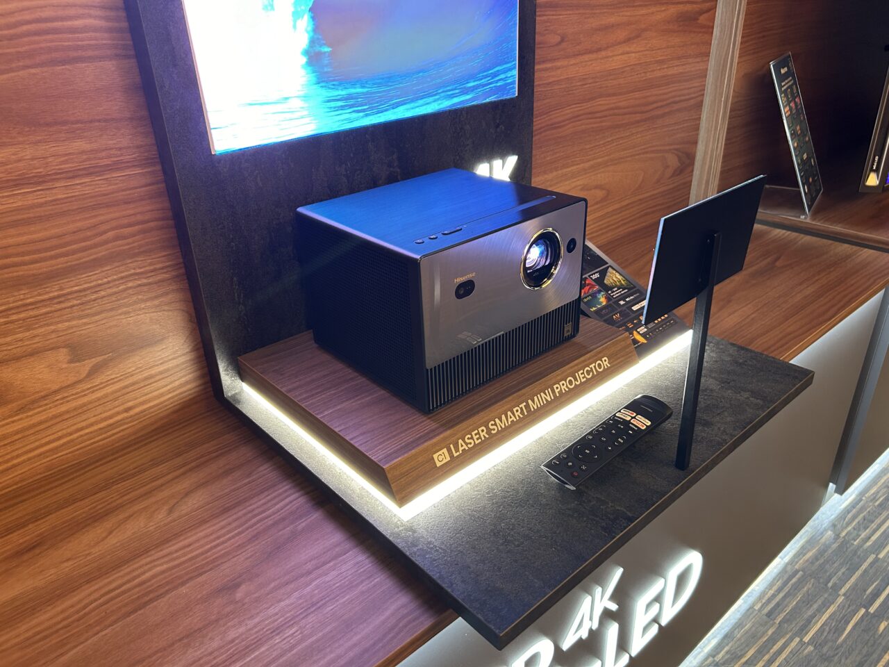 Projektor laserowy smart mini ustawiony na wystawie w sklepie, obok ekran z wyświetlanym obrazem, z pilotem i broszurami informacyjnymi.