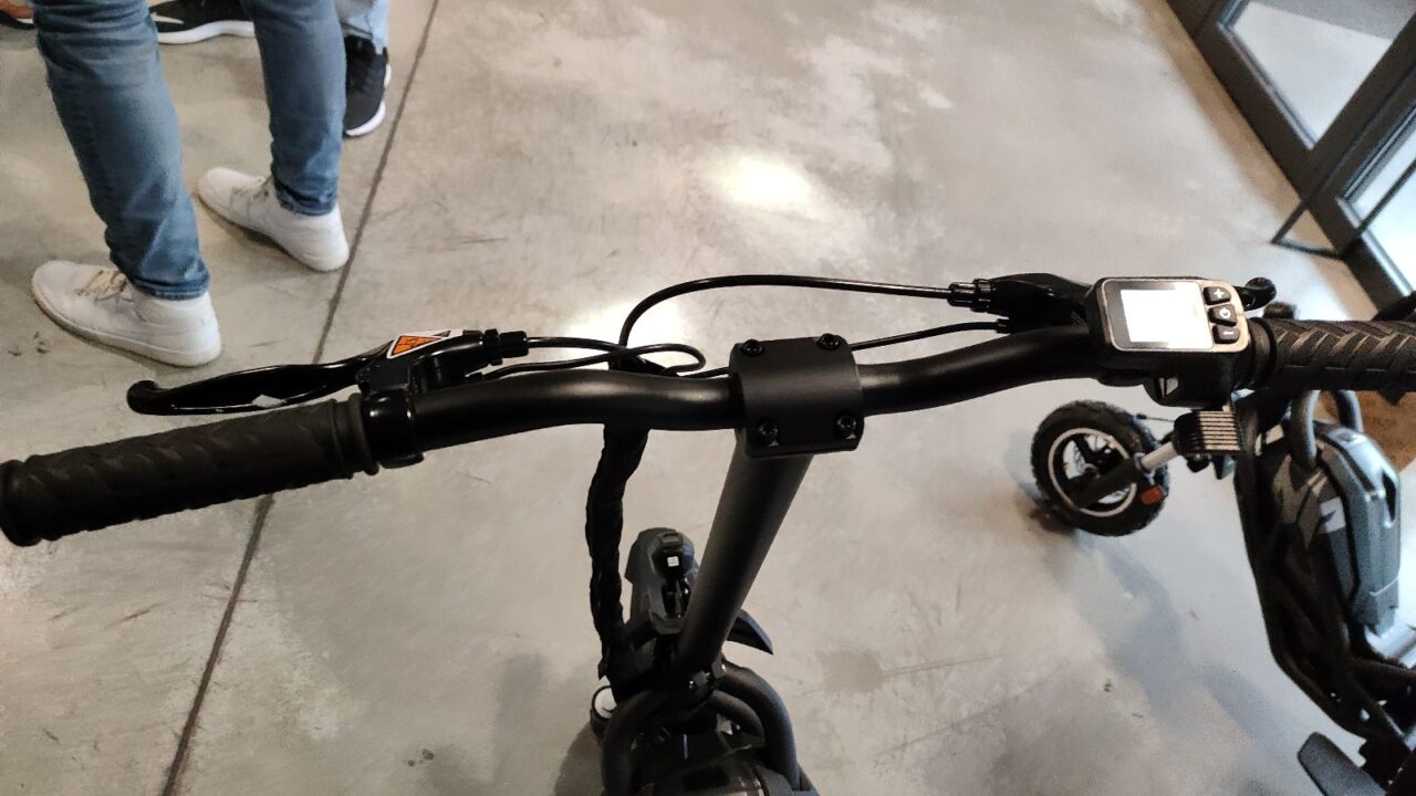 Kierownica roweru z zamocowanym licznikiem rowerowym, w tle niewyraźny fragment roweru dziecięcego i nogi osoby.