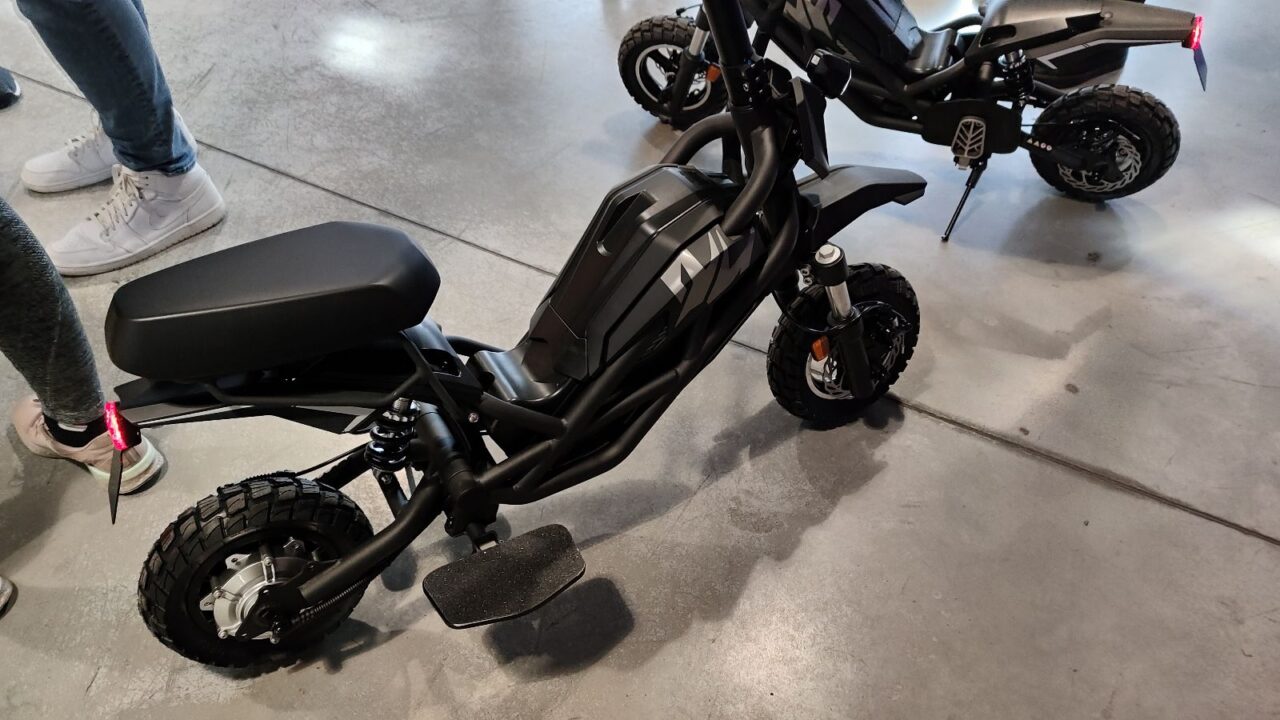 Czarny elektryczny motocykl o terenowym wyglądzie na wystawie, w tle częściowo widoczna noga osoby.