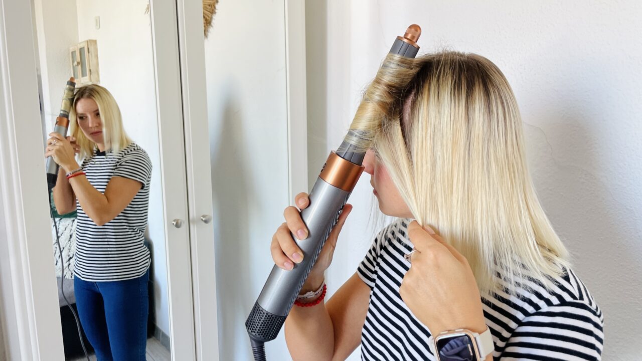 Kobieta używa lokówki do kręcenia włosów przed lustrem w domowym wnętrzu.