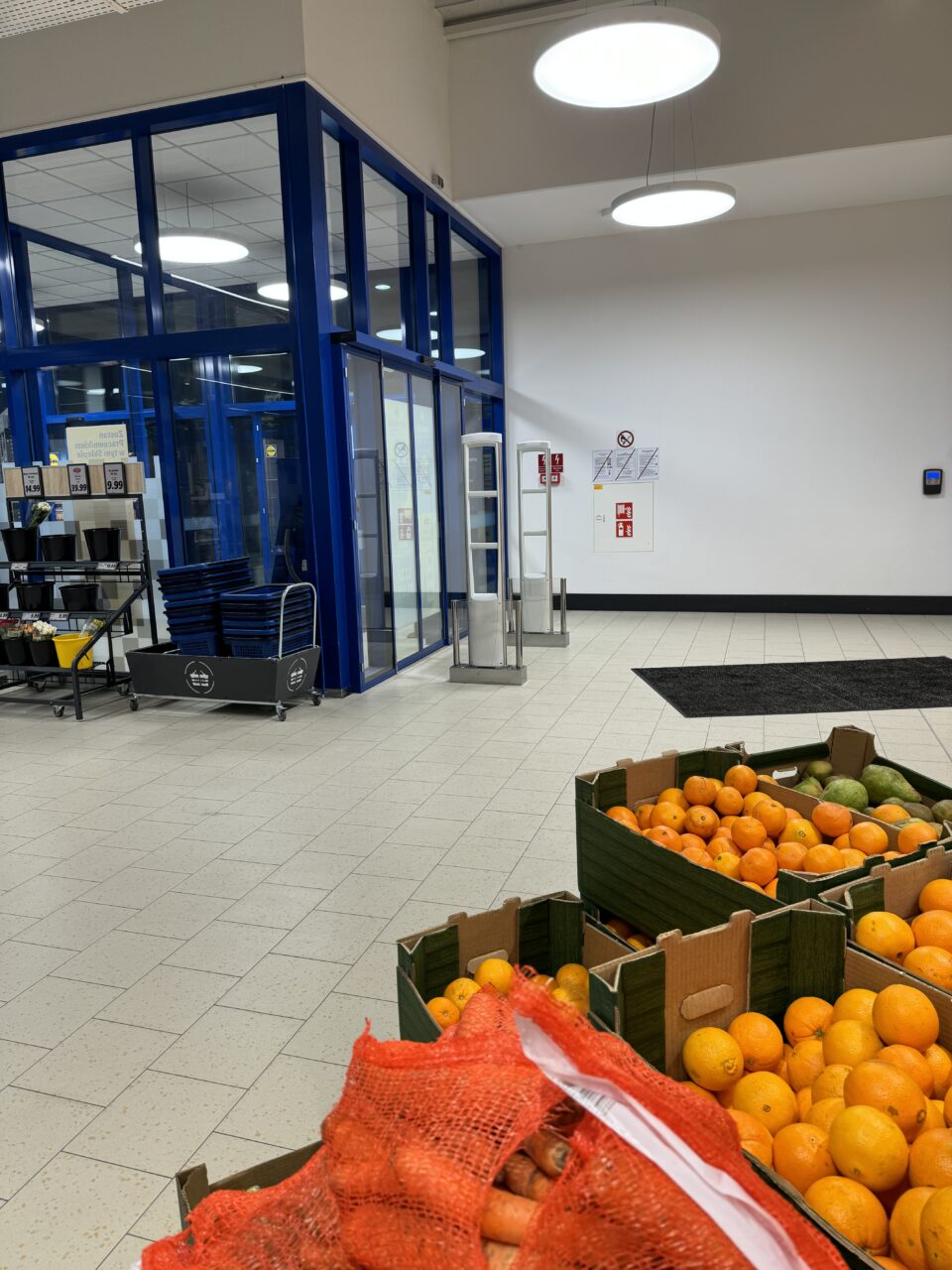 Wejście do supermarketu Lidl z wyeksponowanymi skrzynkami ze świeżymi pomarańczami i marchewką, widoczne wózki na zakupy, niebieskie drzwi i plakaty z instrukcjami bezpieczeństwa.