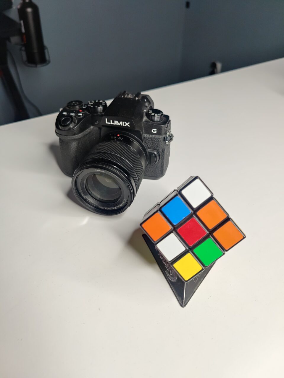 Aparat fotograficzny marki Lumix umieszczony na białej powierzchni w tle, z prawej strony w pierwszym planie częściowo rozwiązana kostka Rubika.