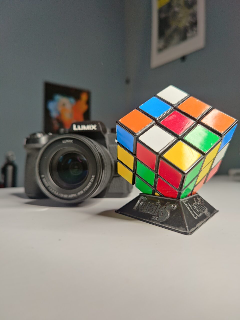 Kostka Rubika na pierwszym planie na czarnym stojaku, z rozmytym aparatem fotograficznym Panasonic Lumix w tle oraz plakatami na ścianie.