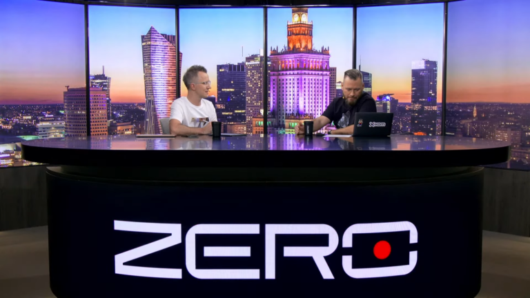 Dwóch mężczyzn siedzi przy biurku z napisem "ZERO" na tle panoramicznego widoku wieczornego miasta.