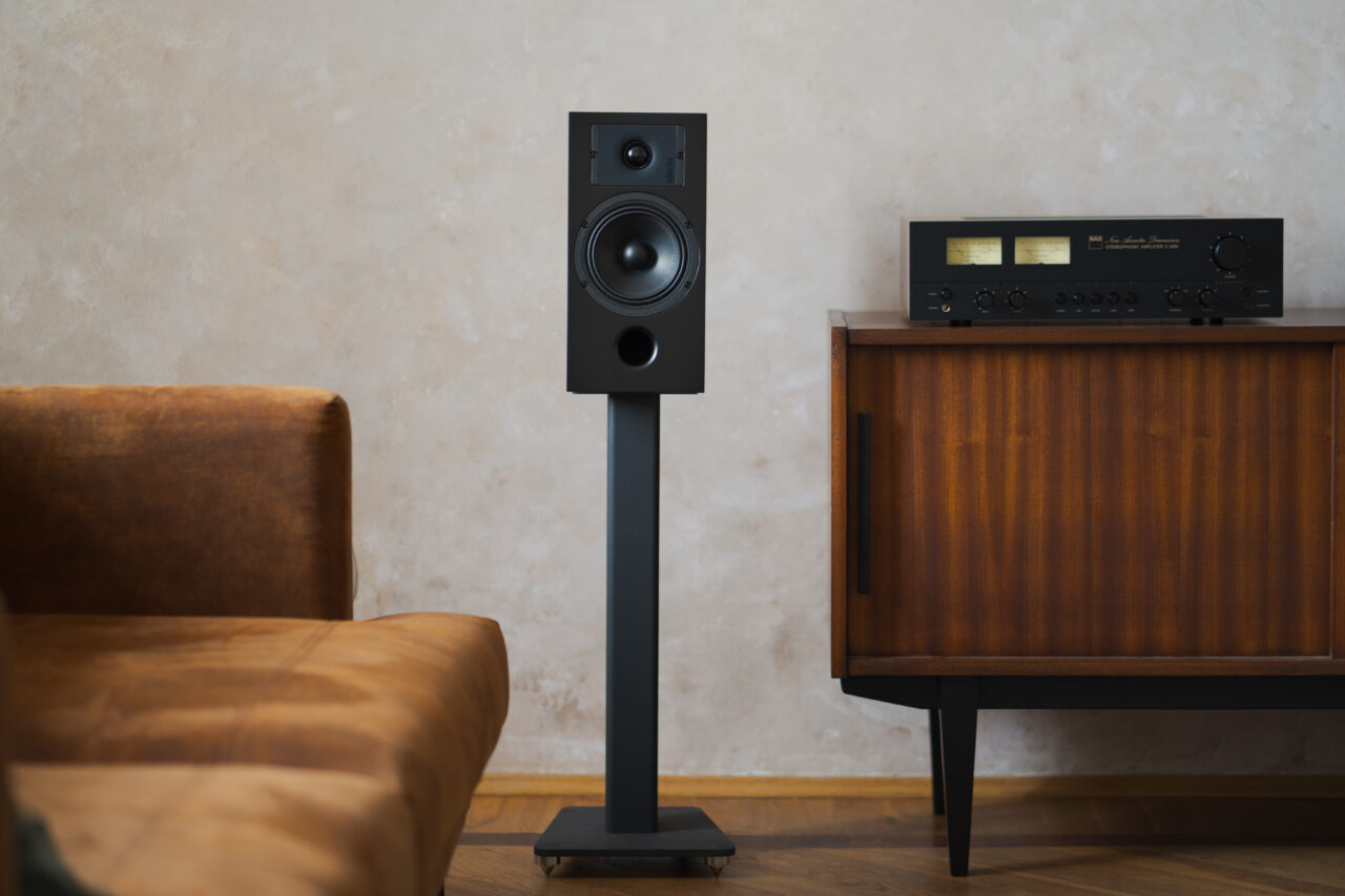 Głośnik na podstawce umieszczony w salonie obok drewnianego mebla z wbudowanym sprzętem audio i brązowej, skórzanej kanapy.