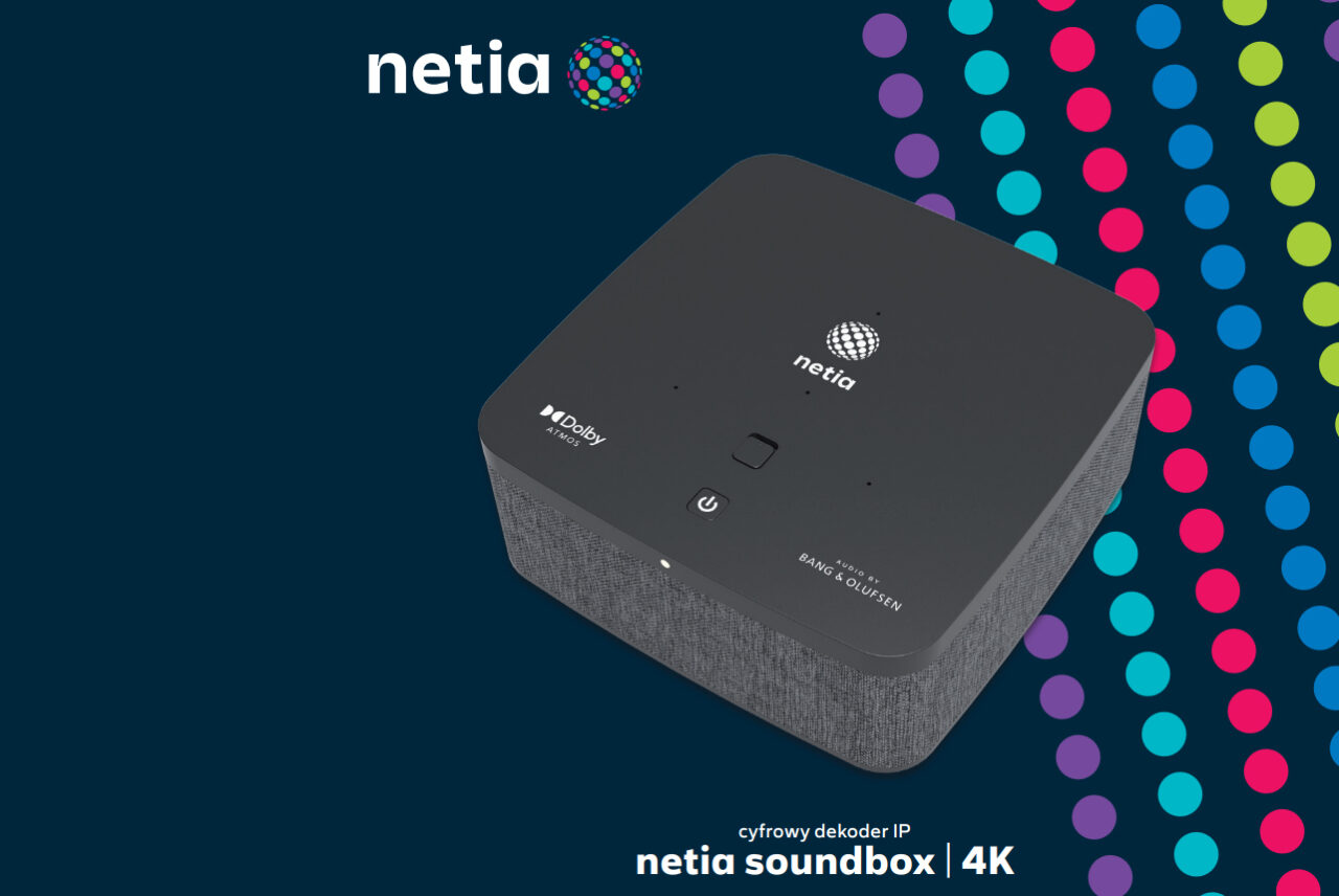 Cyfrowy dekoder  Netia Soundbox 4K na ciemnym tle z kolorowymi kropkami, logo Netia w górnej lewej części obrazu. Dekoder ma szare wykończenie z teksturą przypominającą tkaninę i czarny górny panel z logotypami Dolby Audio i Bang & Olufsen oraz przyciskami zasilania i Bluetooth.