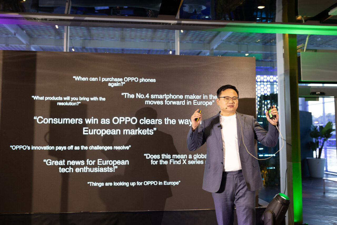 Mężczyzna w garniturze stoi na scenie w czasie prezentacji, w tle duży ekran z angielskimi tekstami na temat firmy OPPO i rynku europejskiego.