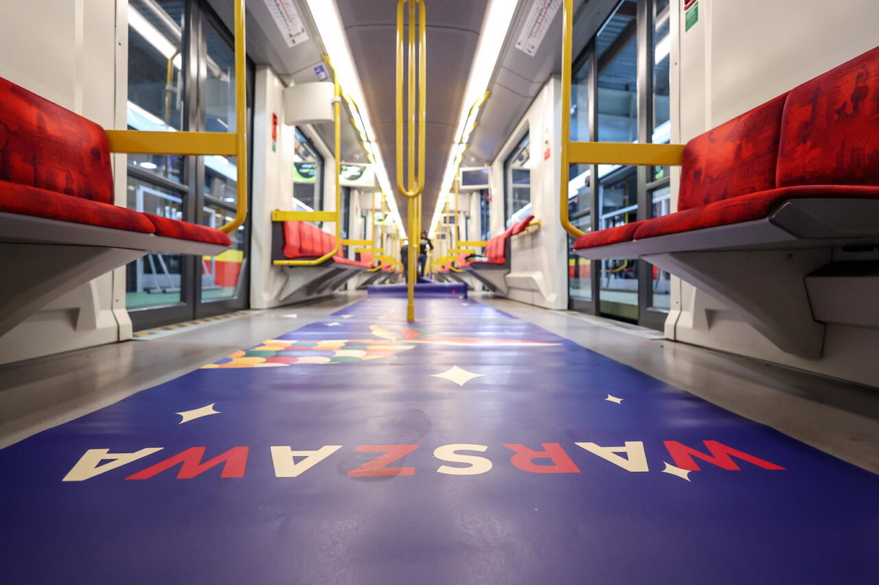 Metro Warszawa. Wnętrze pustego wagonu z czerwonymi siedzeniami i żółtymi poręczami, z podłogą pokrytą wzorami i literami.
