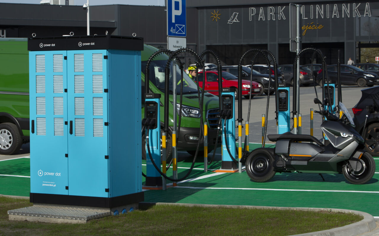 Stacja ładowania pojazdów elektrycznych z wieloma ładowarkami i szafką na ładowarki, hulajnoga elektryczna zaparkowana obok, w tle parking i budynek z napisem "PARK LINIANKA ejście".