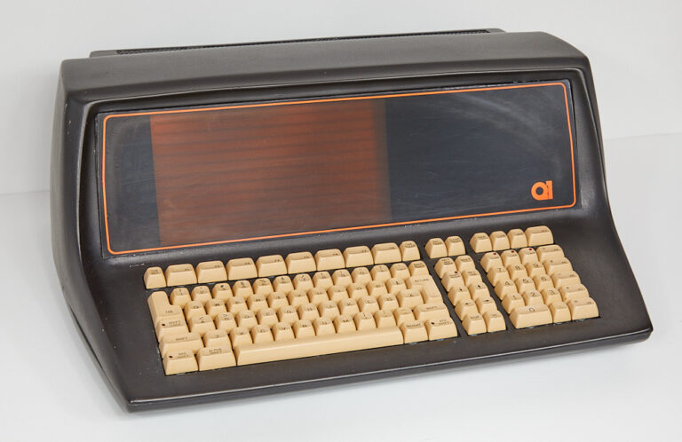 Stary komputer osobisty z klawiaturą w kolorze kości słoniowej i wbudowanym monitorem CRT w obudowie w kolorze czarnym z pomarańczowymi akcentami.