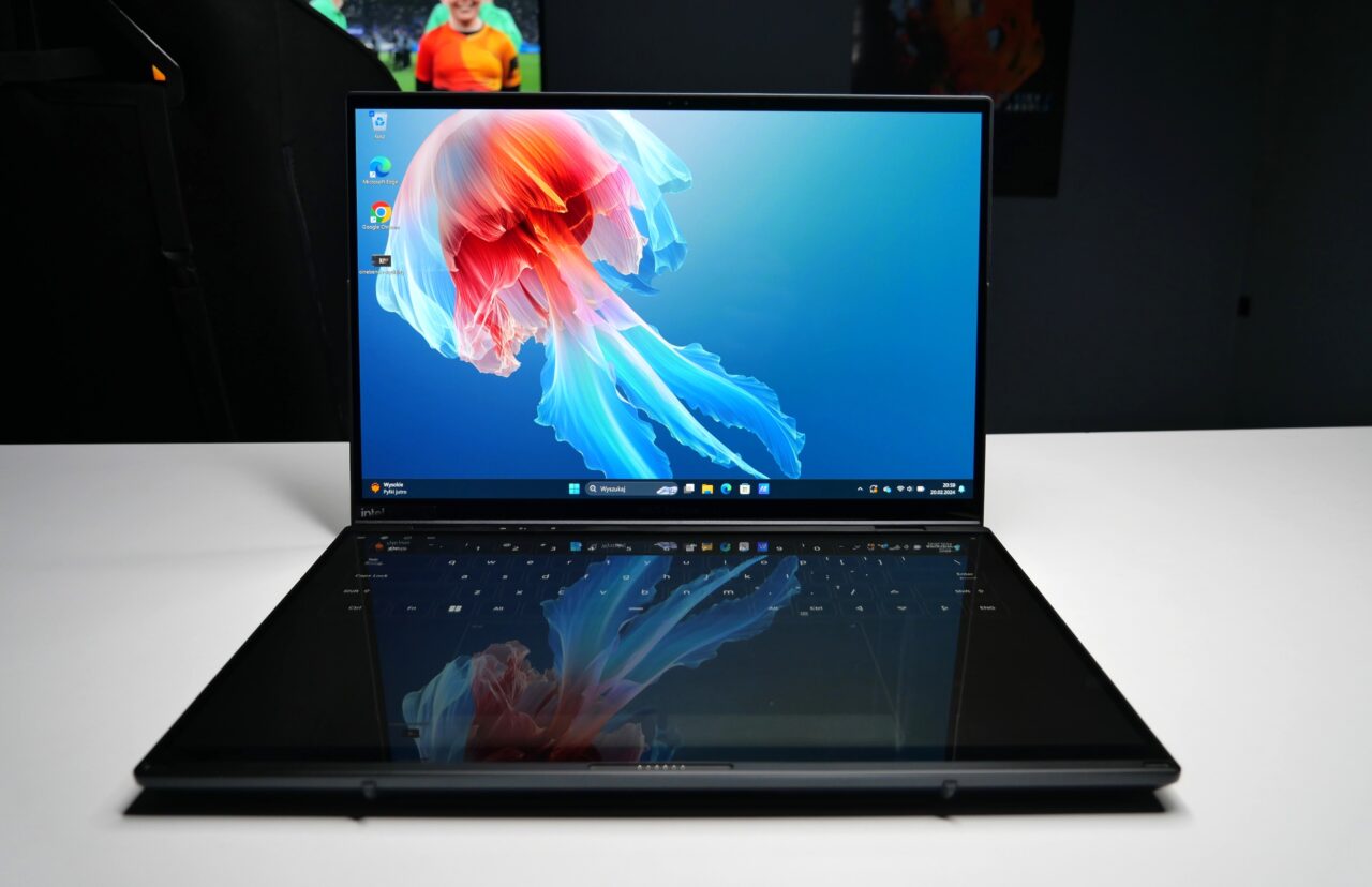 Laptop z włączonym ekranem przedstawiającym obraz meduzy na tle w kolorze błękitnym, ustawiony na białym stole w pomieszczeniu oświetlonym rozproszonym światłem.
