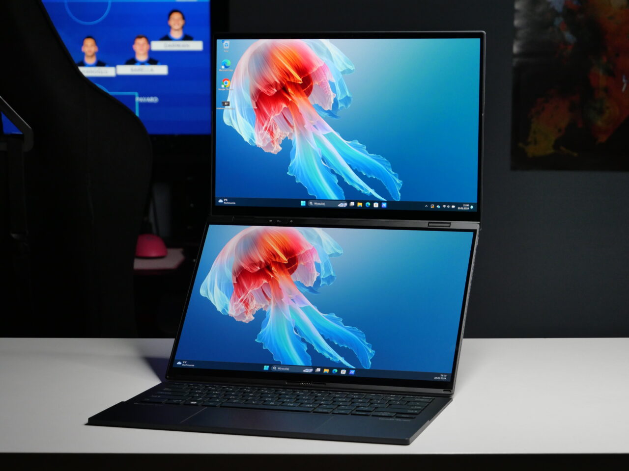 Laptop z dwoma ekranami, oba wyświetlają to samo graficzne tło z abstrakcyjnym wzorem. W tle widoczny fragment biurka z monitorem wyświetlającym interfejs użytkownika.