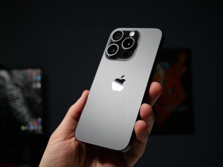 Tylna część srebrnego smartfona z trzema obiektywami aparatu i logo firmy Apple, trzymana w dłoni na tle rozmytego ciemnego pokoju.