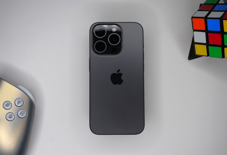 Czarny smartfon Apple iPhone 15 Pro z potrójnym aparatem leży na białym tle obok częściowo widocznej srebrnej konsoli do gier i kolorowej kostki Rubika.