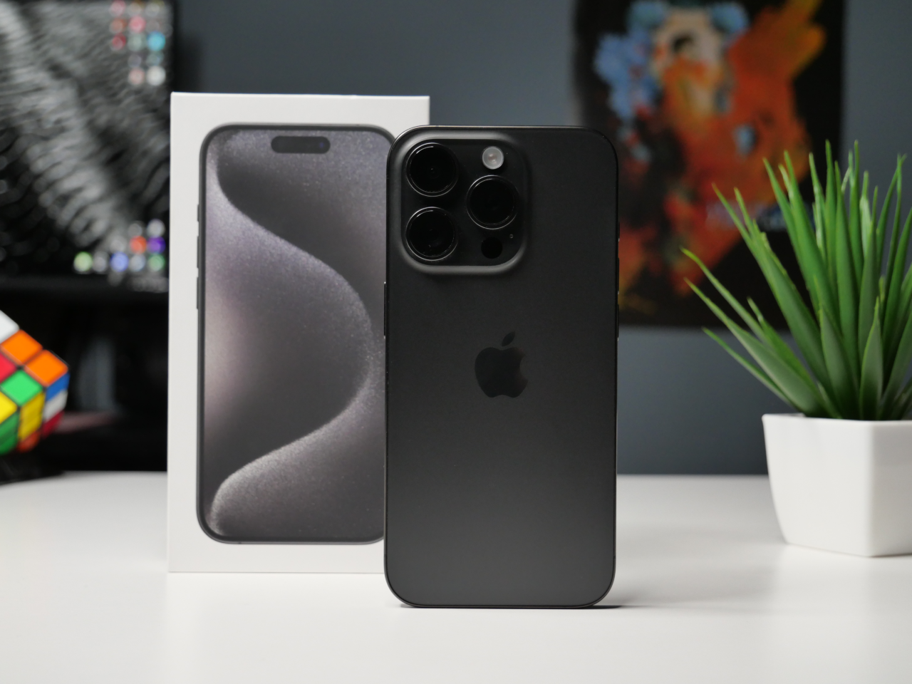 Czarny iPhone z potrójnym aparatem umieszczony obok swojego pudełka, w tle roślina doniczkowa i kostka Rubika na biurku.