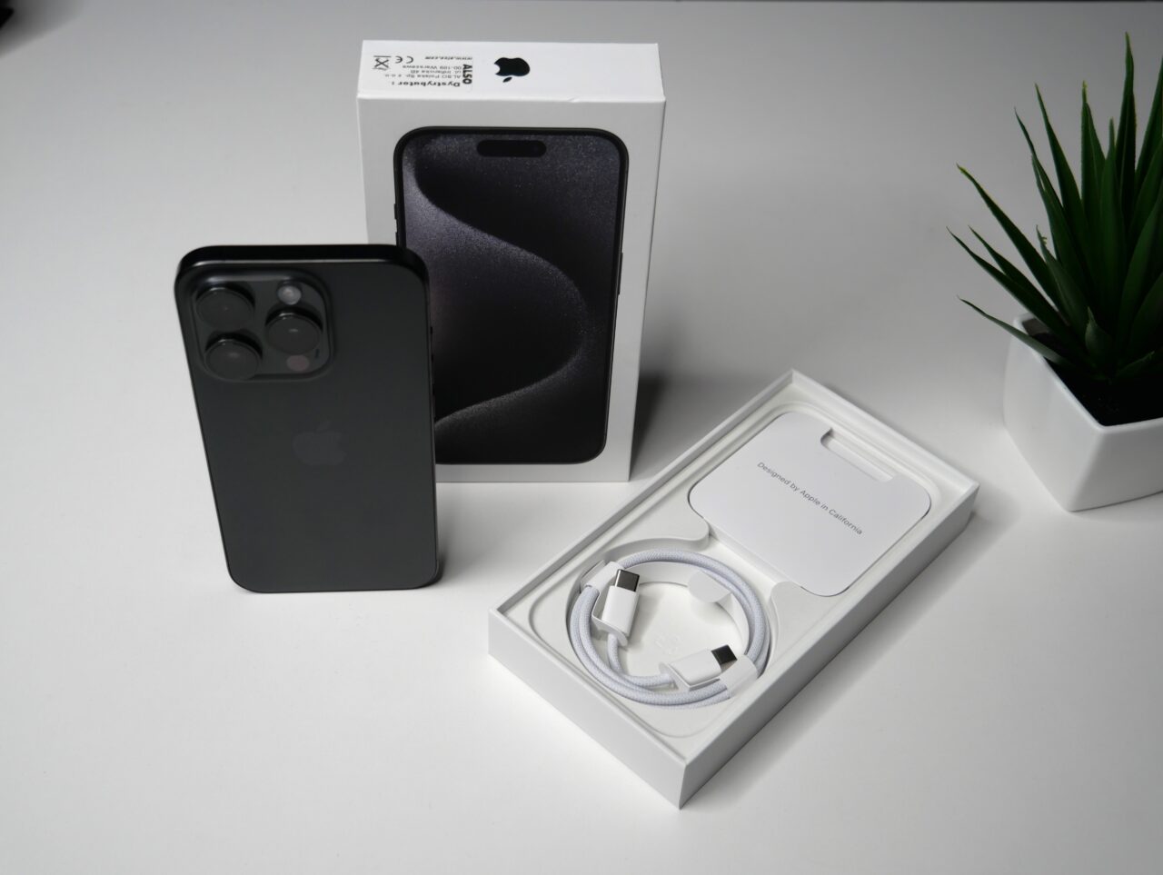 Czarny iPhone z potrójnym aparatem umieszczony obok otwartego pudełka, wewnątrz którego znajduje się kabel do ładowania, przy obecności pudełka od iPhone'a i doniczki z zieloną rośliną.
