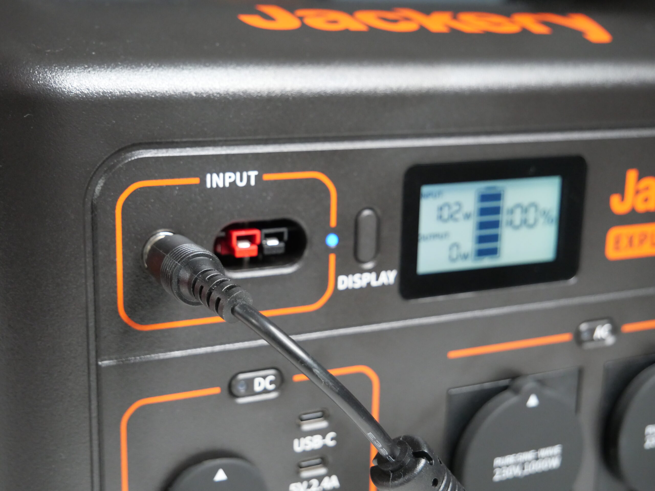 Wtyczka ładowarki podłączona do przenośnego generatora z logo Jackery, z wyświetlaczem wskazującym naładowanie baterii na 100%.