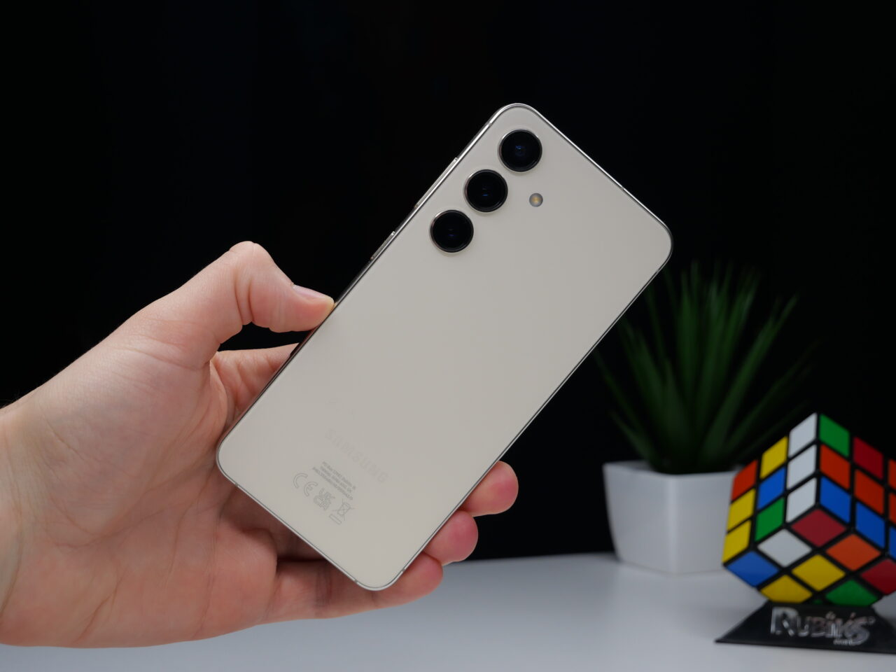 Parte traseira de um smartphone na mão, com câmera tripla e logotipo do fabricante, tendo como fundo uma tela preta, uma planta verde e um cubo de Rubik.