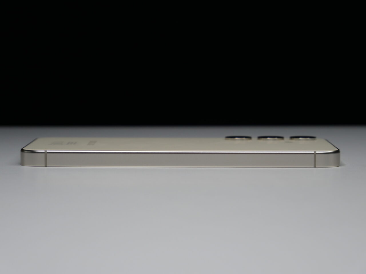Zbliżenie na złoty smartfon leżący bokiem, na białej powierzchni i ciemnym tle, widać przyciski i porty.