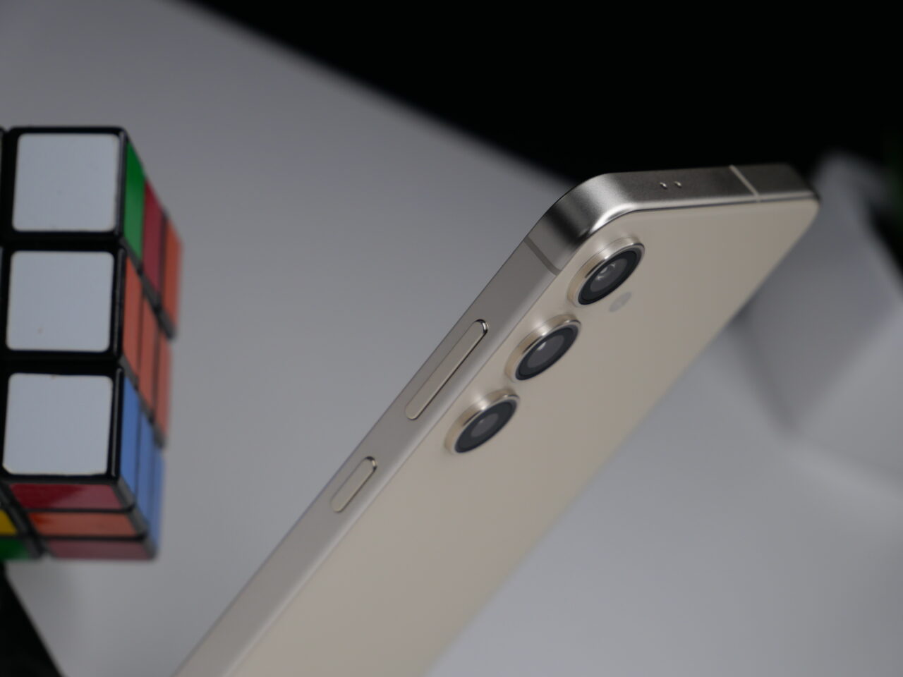 Zbliżenie na złoty smartfon z potrójnym aparatem i przyciskami po lewej stronie, z rozmytym kolorowym kostką Rubika w tle.