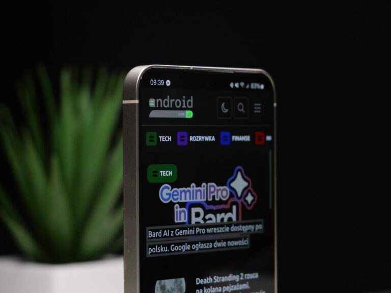 Smartfon Samsung Galaxy S24 wyświetlający stronę internetową z artykułami o tematyce technologicznej, stojący na stole z rozmazanym zielonym roślinnym tłem.