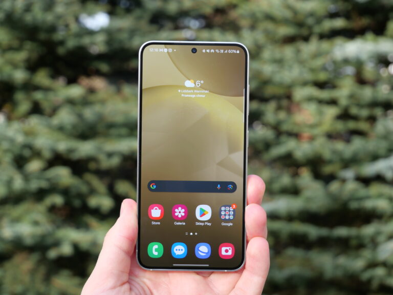 Ręka trzymająca smartfon Samsung Galaxy S24 z ekranem głównym wyświetlającym aplikacje na tle rozmazanych zielonych krzewów.