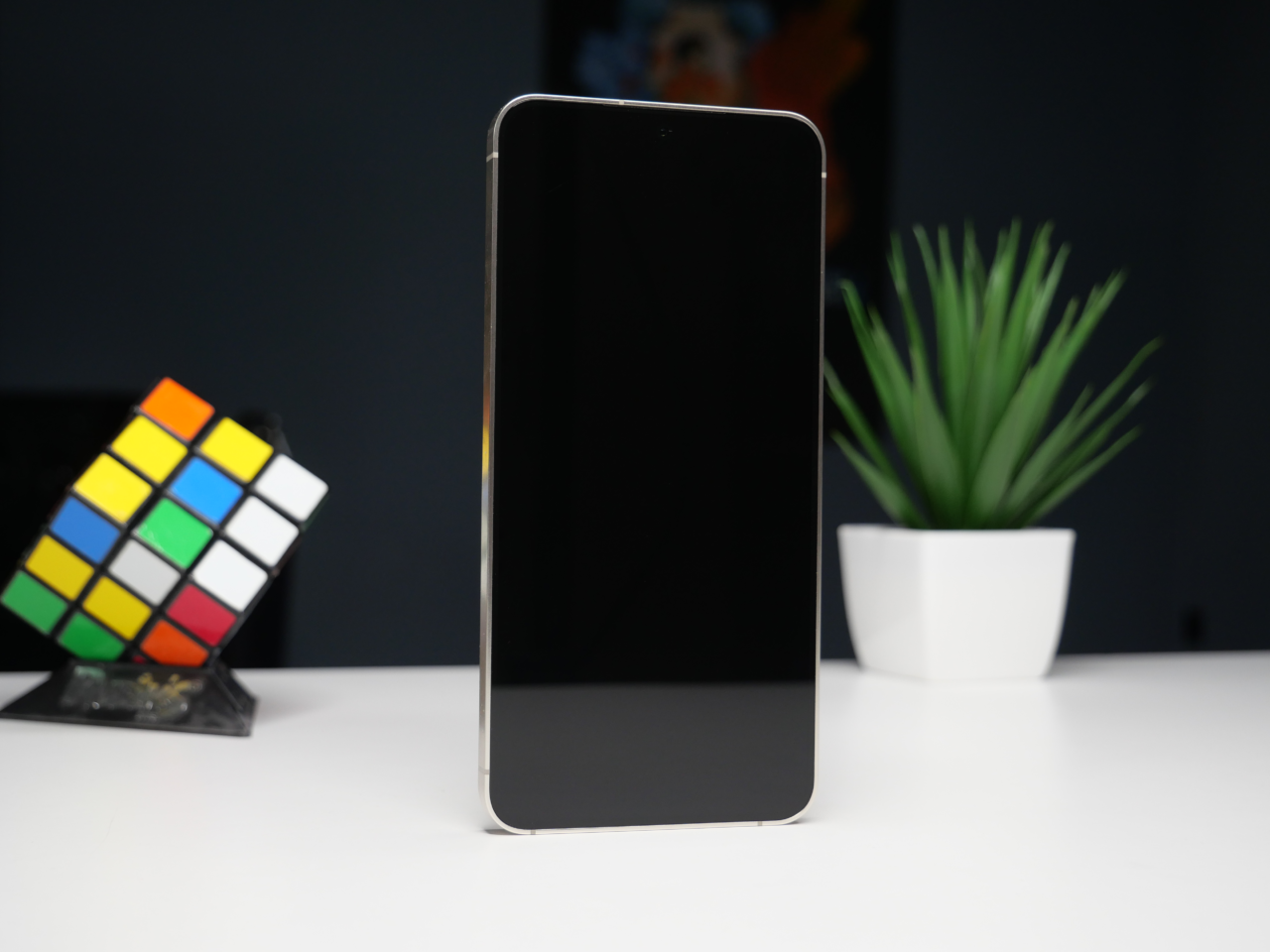 Smartfon stoi pionowo na biurku, wyłączony ekran skierowany do przodu; w tle roślina doniczkowa i kostka Rubika.
