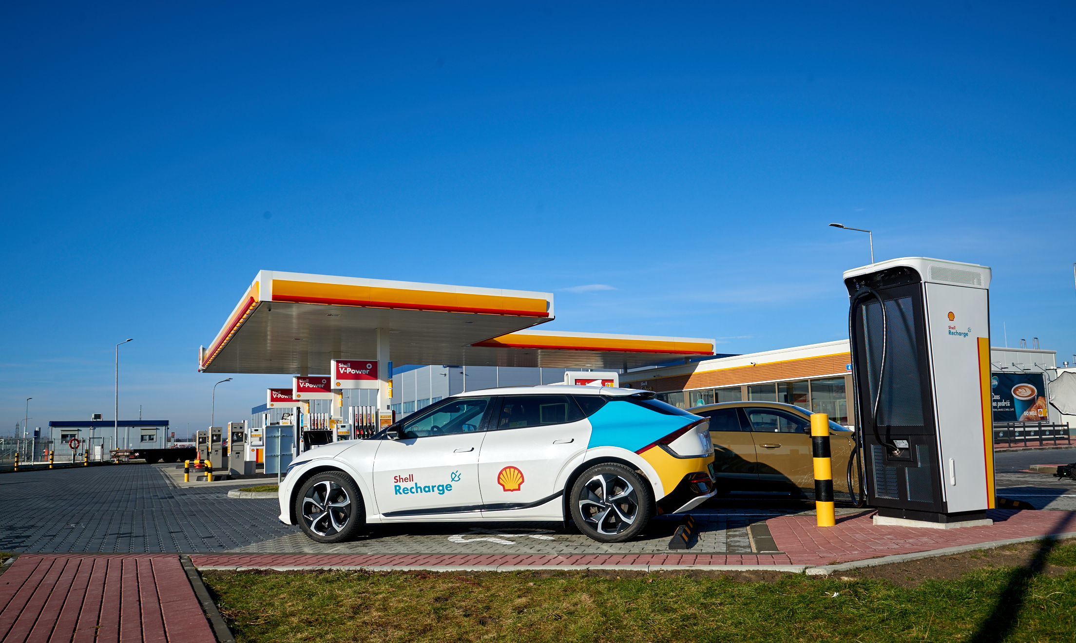 Elektryczny samochód podłączony do stacji ładowania na stacji benzynowej Shell w słoneczny dzień.