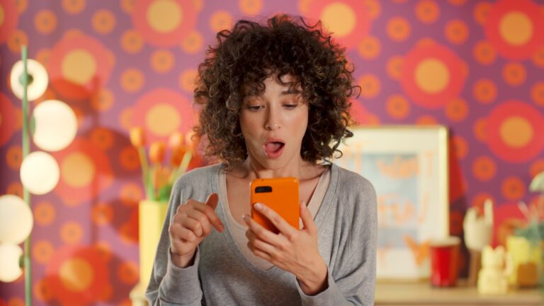 Zaskoczona kobieta z kręconymi włosami patrzy na telefon komórkowy na tle kolorowej, wzorzystej ściany.