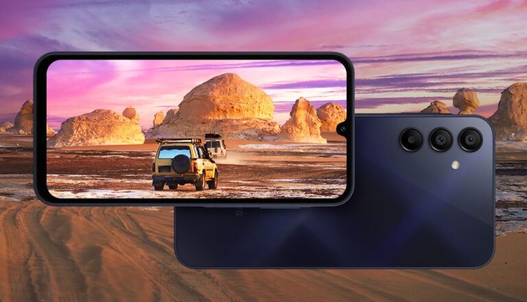 Smartfon Samsung Galaxy A15 leżący na powierzchni z widokiem pustyni i jadącym samochodem terenowym wyświetlanym na ekranie, z tyłu aparat fotograficzny z trzema obiektywami.