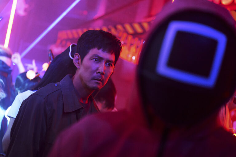 Drugi sezon Squid Game. Mężczyzna patrzy zatroskany przez tłum na imprezie z neonowym oświetleniem, przed nim osoba w masce z kwadratowym, świecącym konturem.