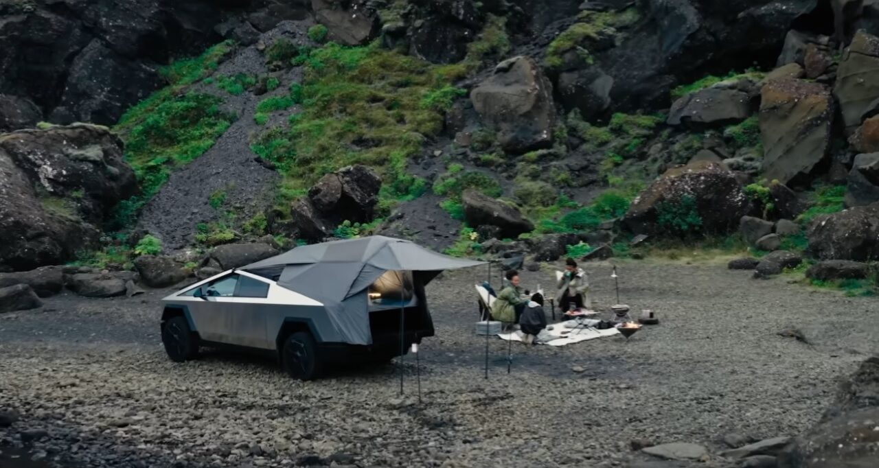 Tesla Cybertruck. Grupa osób wypoczywa na biwaku w górskiej, skalistej okolicy, obok stojącego futurystycznego samochodu z namiotem dachowym.