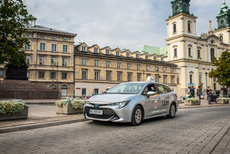 Srebrny samochód marki Toyota z oznaczeniami Uber jako taksówka na tle zabytkowych kamienic i kościoła w jasnych barwach w mieście.