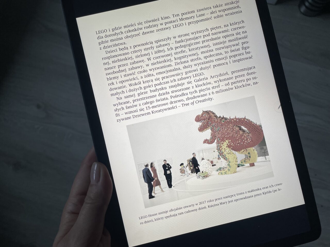 Osoba przygląda się ekranowi tabletu, na którym wyświetlony jest artykuł z fotografiami. Na głównym zdjęciu widać dużą figurę dinozaura z klocków LEGO i grupę ludzi przyglądających się wystawie.