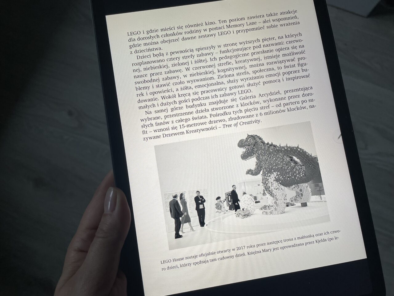 Zdjęcie ekranu urządzenia elektronicznego wyświetlającego artykuł z tekstem w języku polskim i czarno-białą fotograficzne przedstawiającą dużą statuę dinozaura zbudowaną z klocków LEGO, z dwoma sylwetkami ludzi patrzącymi na nią w tle.