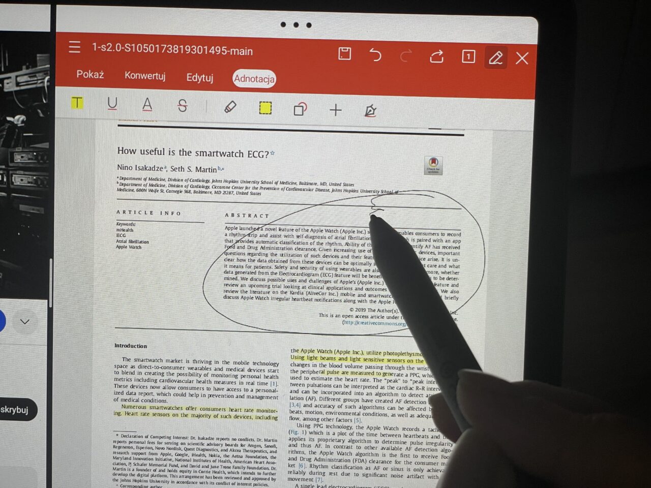 Dłoń używająca cyfrowego pióra do podkreślenia tekst na ekranie tabletu wyświetlającego naukowy artykuł zatytułowany "How useful is the smartwatch ECG?".