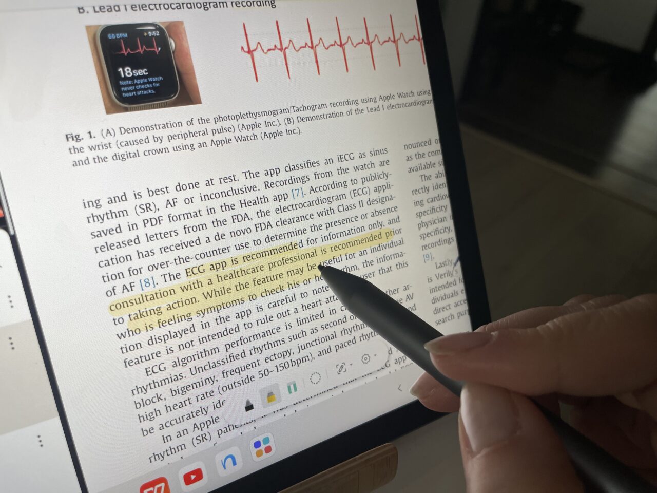 Osoba trzymająca długopis czyta naukowy artykuł na tablecie, na którym widać wykres elektrokardiogramu oraz fragment tekstu o aplikacji EKG w zegarku Apple Watch.