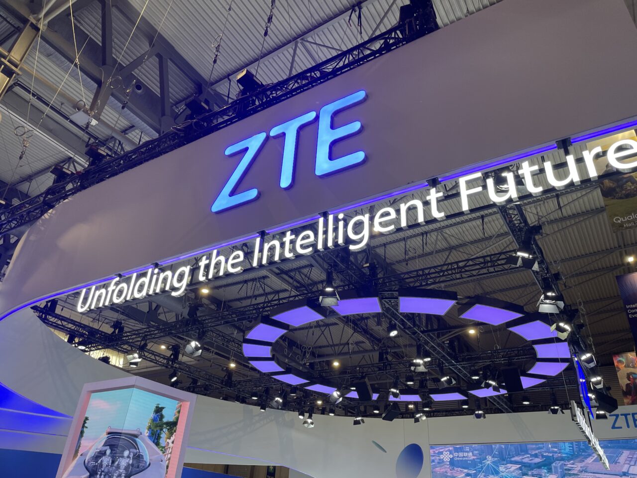 Zdjęcie stoiska firmy ZTE z niebieskimi literami na górze i hasłem "Unfolding the Intelligent Future" na tle wystawy technologicznej.