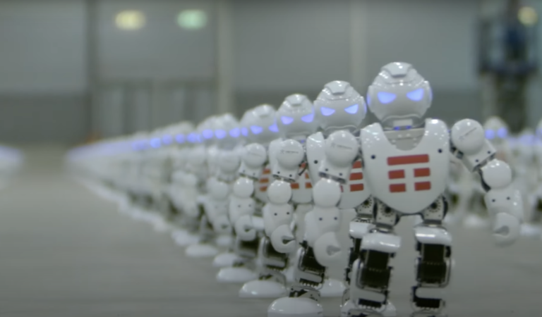 Szereg małych, białych robotów humanoidalnych z niebiesko świecącymi oczami wewnątrz pomieszczenia.