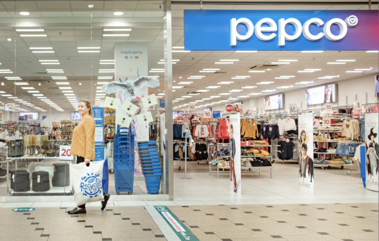 Kobieta opuszcza sklep Pepco z torbą na zakupy, przeszklone sklepowe witryny, w tle rozmieszczone są różne produkty odzieżowe, na pierwszym planie kilka stojaków i reklam.