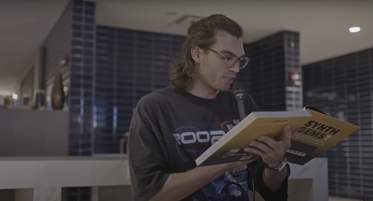 Mężczyzna z długimi włosami i okularami czyta książkę zatytułowaną "Synth Gems" stojąc w pomieszczeniu z ciemnymi szafami w tle.