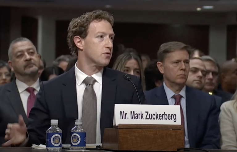 Mark Zuckerberg w garniturze siedzący przed mikrofonem z tabliczką z napisem "Mr. Mark Zuckerberg", w tle grupa ludzi.